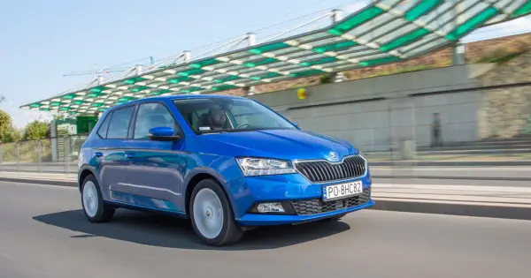 Niebieska Škoda Fabia jedzie po drodze, za nią widać nowoczesną konstrukcję z zielonym zadaszeniem.