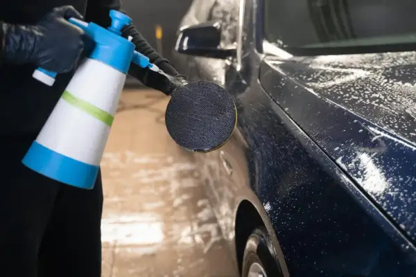 En person med svarta handskar sprutar skum på en mörk metallicbil med en blå och vit sprayflaska. Bilvax metallic ger en glansig finish.