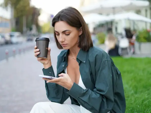 Kobieta z oliwkowym kolorem skóry, trzymająca kawę i telefon, siedzi na ławce w mieście.