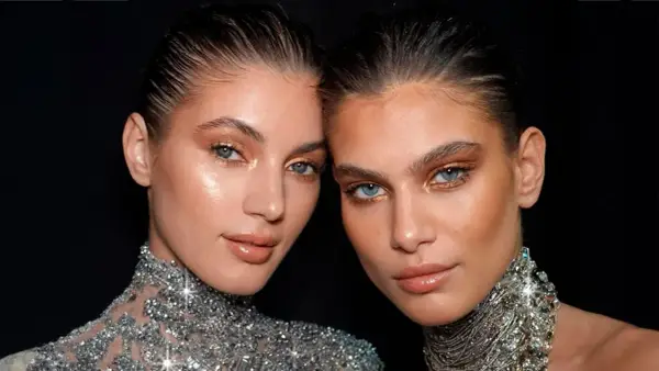 Dos mujeres con maquillaje dorado y un sutil rabillo del ojo, luciendo collares brillantes.