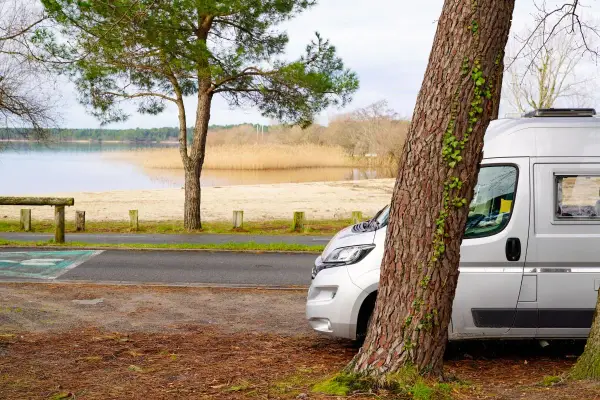 Un fourgon de 6 mètres est garé près d'un lac, à l'ombre d'un grand arbre. Le paysage est paisible, avec une plage de sable et des roseaux au loin.