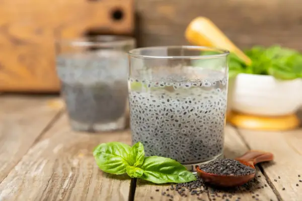 Due bicchieri con semi di basilico in acqua, un cucchiaino di semi e foglie fresche su un tavolo di legno.