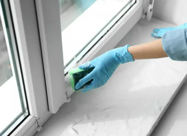 Ręka w niebieskiej rękawiczce czyści parapet gąbką. Dowiedz się, jak usunąć klej po taśmie z drzwi i innych powierzchni.