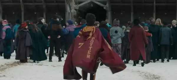 Chłopiec w pelerynie z napisem "POTTER 7" idzie przez śnieg w otoczeniu innych uczniów, jak w serialu Harry Potter.