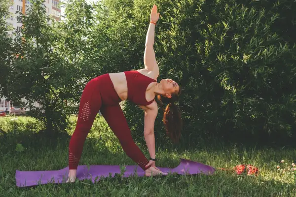 Kobieta w czerwonym stroju wykonuje pozycję Trójkąta podczas praktyki jogi ashtanga na trawie, otoczona zielenią.