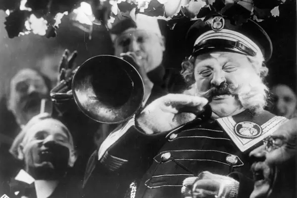 Un uomo con baffi e barba folta suona una tromba, circondato da volti sorridenti. Un'atmosfera da kammerspiel, un dramma intimo e intenso.