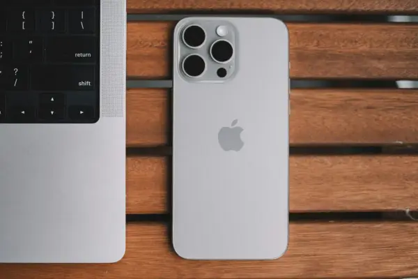Srebrny iPhone 13 Pro z systemem iOS 26.4 obok klawiatury MacBook'a na drewnianym stole.