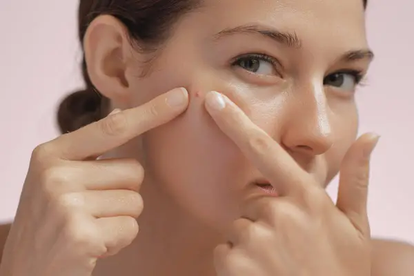 Une femme examine un furoncle sur son visage, ses doigts pointant vers la petite imperfection rouge sur sa joue.