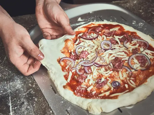 Hände ziehen den rohen Pizzateig vom Pizzaschieber. Der Teig klebt leicht am Schieber, während die Pizza mit Tomatensauce, Käse, Salami und Zwiebeln belegt ist.
