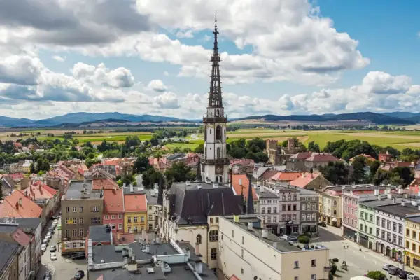Urokliwe miasto na trasie Wrocław, z dominującą wieżą ratuszową i kolorowymi kamienicami, otoczone zielonymi polami i wzgórzami.