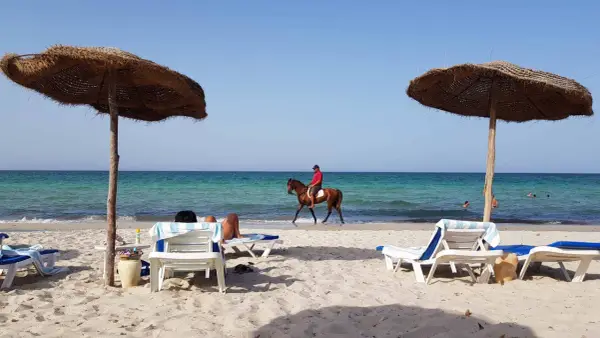 Piękna plaża na Djerbie, Tunezja. Jeździec na koniu przemierza brzeg morza, podczas gdy inni odpoczywają pod parasolami.