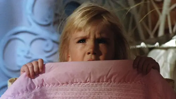 Bambina spaventata si nasconde sotto una coperta rosa, gli occhi sgranati in un'espressione di puro horror psicologico.
