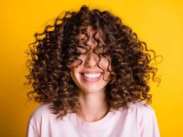 Mujer sonriendo con cabello rizado castaño sobre fondo amarillo.