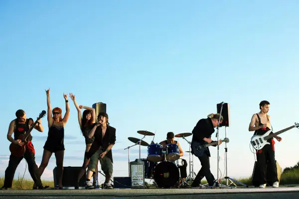 Zespół rockowy na tle nieba. Wokalista, gitarzysta, basista, perkusista i dwie tancerki. Zespoły rockowe zagraniczne alfabetycznie.