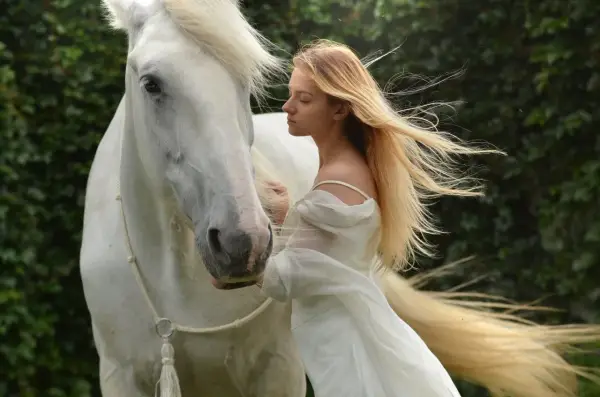 Una joven rubia con un vestido blanco se inclina hacia un majestuoso caballo blanco. Es como soñar con caballos, una escena etérea y serena.