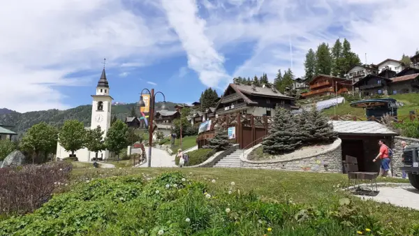 A picturesque village in Chamois, Italy, with a church tower, wooden chalets, and lush greenery. Scopri cosa vedere in questo incantevole borgo alpino.