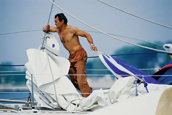 Un marinier, peut-être Eric Tabarly, manipule des voiles sur un bateau. Sa fortune est dans le vent et l'océan.