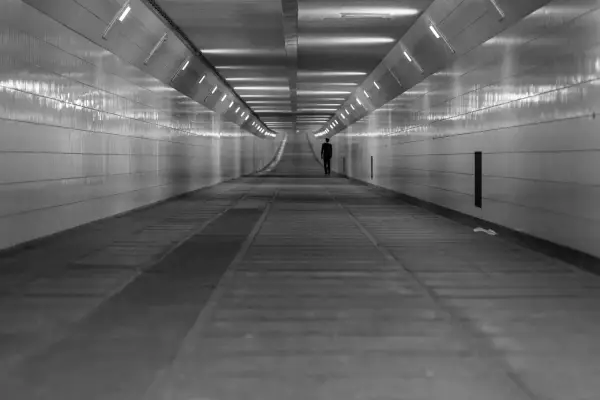 Człowiek w garniturze idzie przez długi, pusty tunel. Światła na ścianach tworzą wrażenie nieskończoności, jakby wchodził w szarą strefę.