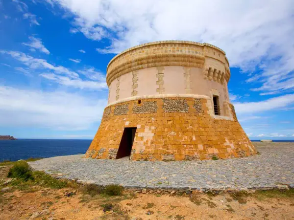 Torre de defensa costera, un lugar histórico que ver en Fornells. El mar azul y el cielo nublado completan la vista.