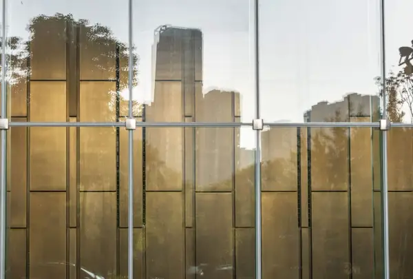 Reflejos dorados de árboles y edificios en un **cristal blindado** moderno, con estructura metálica visible.