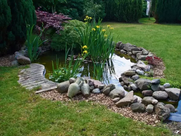 Urokliwe oczko wodne w ogrodzie, otoczone kamieniami i zielenią. Woda odbija niebo, a obok stoją stare amfory.