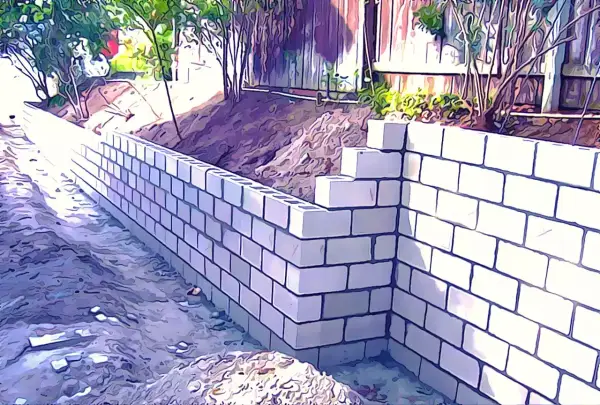 Un muro de contención de bloques de hormigón se está construyendo en un jardín, con tierra y vegetación detrás.