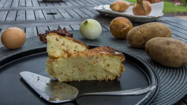 Babka ziemniaczana przepis babci: złocista, pachnąca babka ziemniaczana z boczkiem, obok surowe ziemniaki, cebula i jajko.
