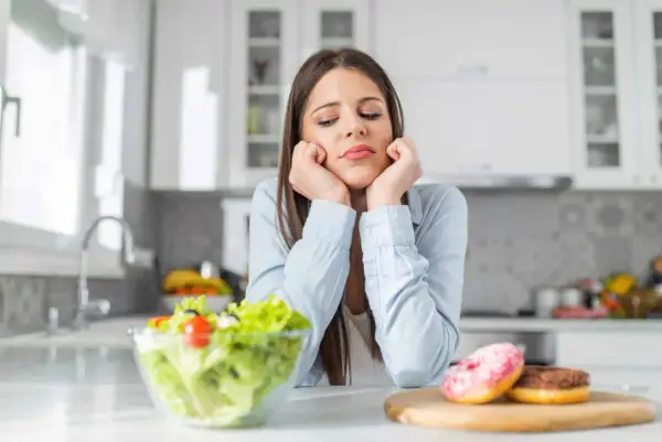 Femme triste face à une salade et des donuts, pensant aux aliments à éviter pour la dermite séborrhéique.