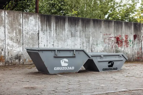Dwa szare kontenery na śmieci zmieszane, z logo GRUZOJAD, stoją przed zniszczonym betonowym murem pokrytym graffiti.