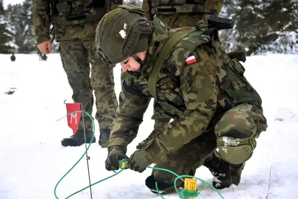Saper w wojsku przygotowuje teren w zimowych warunkach. Żołnierz w kamuflażu pochyla się nad zielonymi przewodami, pracując ostrożnie.