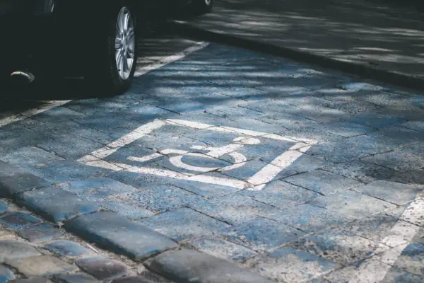 Niebieskie miejsce parkingowe dla niepełnosprawnych pod blokiem, z symbolem wózka inwalidzkiego. Obok widoczna część czarnego samochodu.