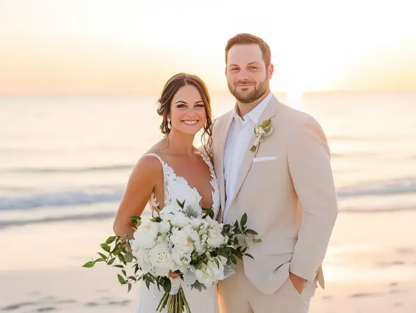 Groom in a tan suit, perfect for what to wear to beach wedding male, smiles with bride holding bouquet at sunset.