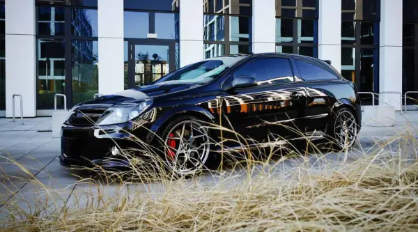 Czarny Opel Astra GTC z chromowanymi felgami i czerwonymi zaciskami hamulcowymi, zaparkowany przed nowoczesnym budynkiem, otoczony suchą trawą.