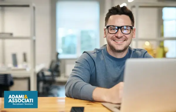 Uomo sorridente con occhiali, seduto alla scrivania con un laptop. Sembra un plant manager che gestisce il suo team.