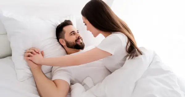 Para trzyma się za ręce w łóżku, patrząc sobie w oczy. Ich spojrzenia pełne są intymności i bliskości.