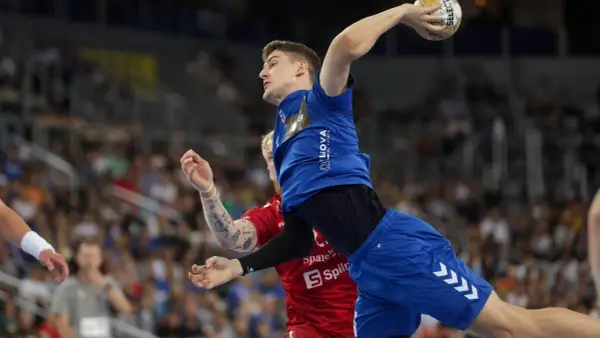 Magnus Landin Jacobsen w skoku do rzutu piłką ręczną, w niebieskim stroju, z widownią w tle.