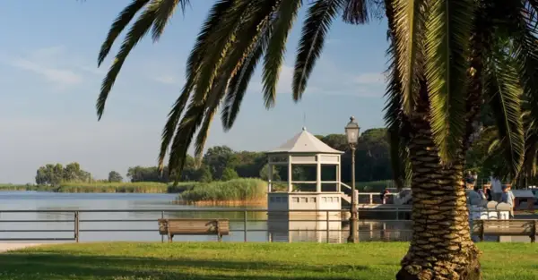 Un gazebo bianco si affaccia sul lago, vicino a una torre. Palme e panchine completano la scena del centro storico.