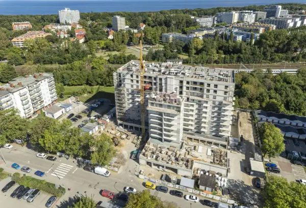 Budowa bloku mieszkalnego w Kołobrzegu. Widok z lotu ptaka na plac budowy, dźwig, samochody i okoliczne budynki. Ceny mieszkań Kołobrzeg mogą być różne.