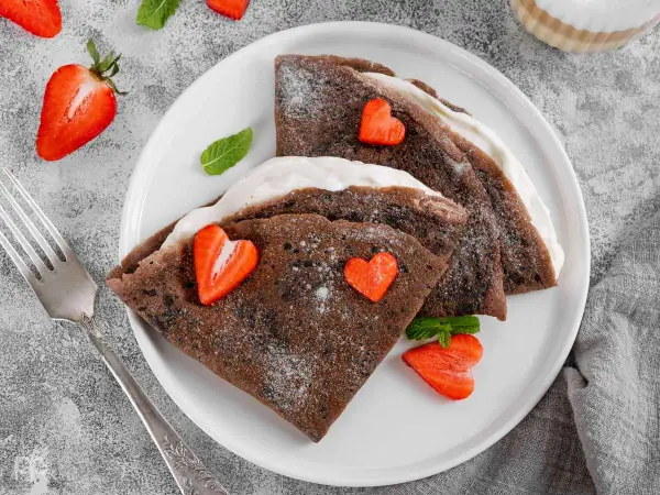 Deliciosos crepes de chocolate rellenos de crema, decorados con fresas en forma de corazón y espolvoreados con azúcar glas.