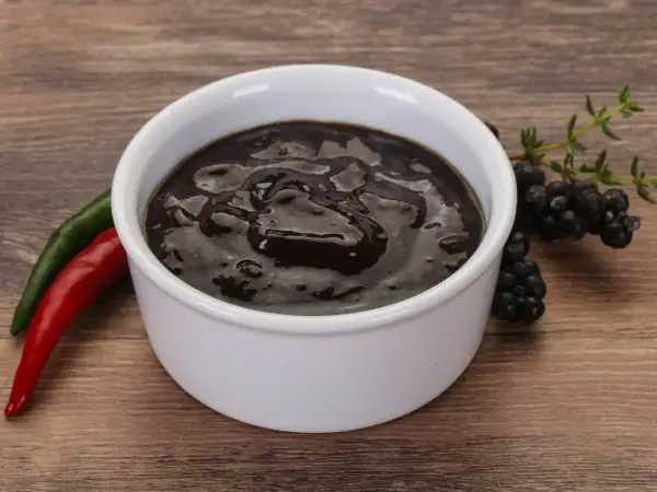 Gęsty, ciemny sos pieprzowy w białej miseczce, obok czerwona i zielona papryczka chili oraz gałązka jeżyn.