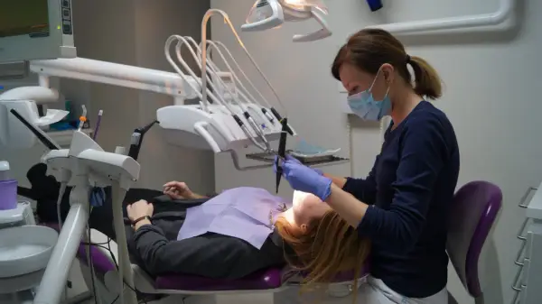 Dentystka w masce i rękawiczkach pracuje przy pacjencie. W gabinecie widoczny sprzęt do stomatologii.