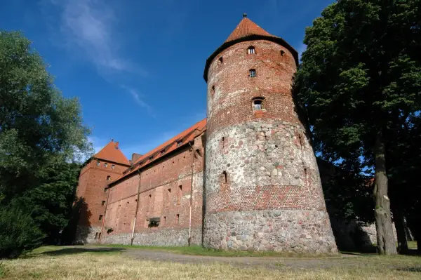 Zamek w Bytowie, potężna ceglana twierdza z kamienną podstawą wieży, otoczona zielenią pod błękitnym niebem.