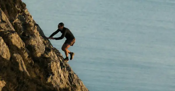Kilian Jornet escalade une paroi rocheuse abrupte, dominant un plan d'eau calme, rappelant l'esprit d'aventure dans les Pyrénées.