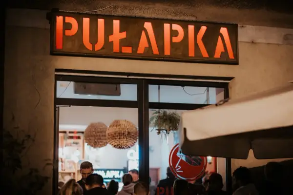 Neonowy napis "PUŁAPKA" nad wejściem do lokalu. W środku widać ludzi i dekoracje.