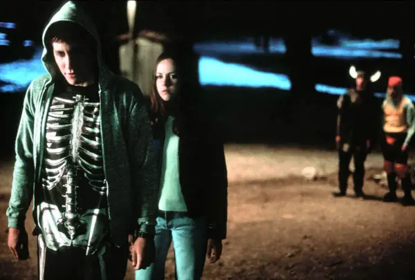Donnie Darko w bluzie z kapturem i koszulce z nadrukiem szkieletu, obok dziewczyny. W tle dwie postacie w kostiumach.