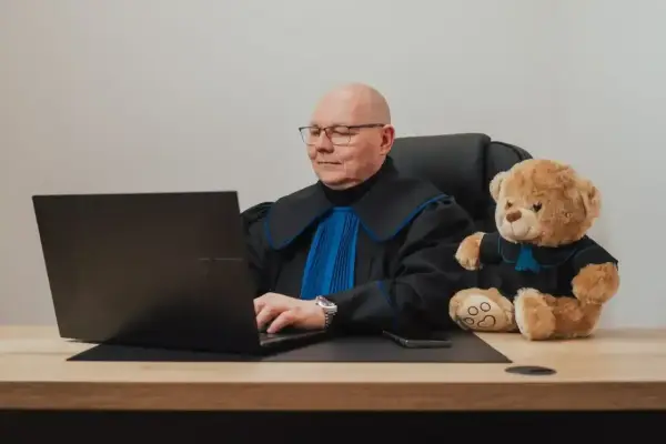 Prawnik w todze z pluszowym misiem obok, pracujący przy laptopie. Zastanawiasz się, ile kosztuje porada prawna?