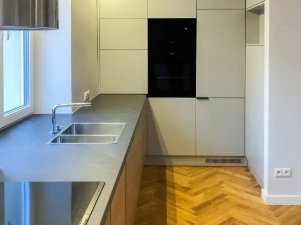 Nowoczesna kuchnia z szarymi frontami, wbudowanym piekarnikiem i podwójnym zlewozmywakiem. Podłoga w jodełkę i okap w kolorze RAL 9016 dodają elegancji.