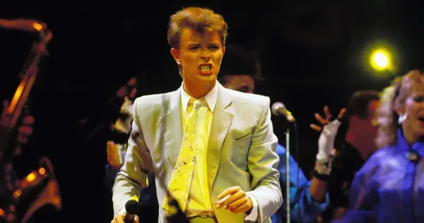 David Bowie na scenie podczas Live Aid, w jasnej marynarce i żółtym krawacie, śpiewa z pasją.