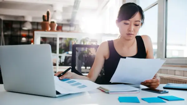 Junge Frau prüft Dokumente, vielleicht die Urlaubsbescheinigung des letzten Arbeitgebers, am Schreibtisch mit Laptop.