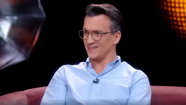 Michał Czernecki w jasnej koszuli i okularach, uśmiecha się. W tle widać fragmenty scenografii, przypominające o jego filmach, serialach i programach.